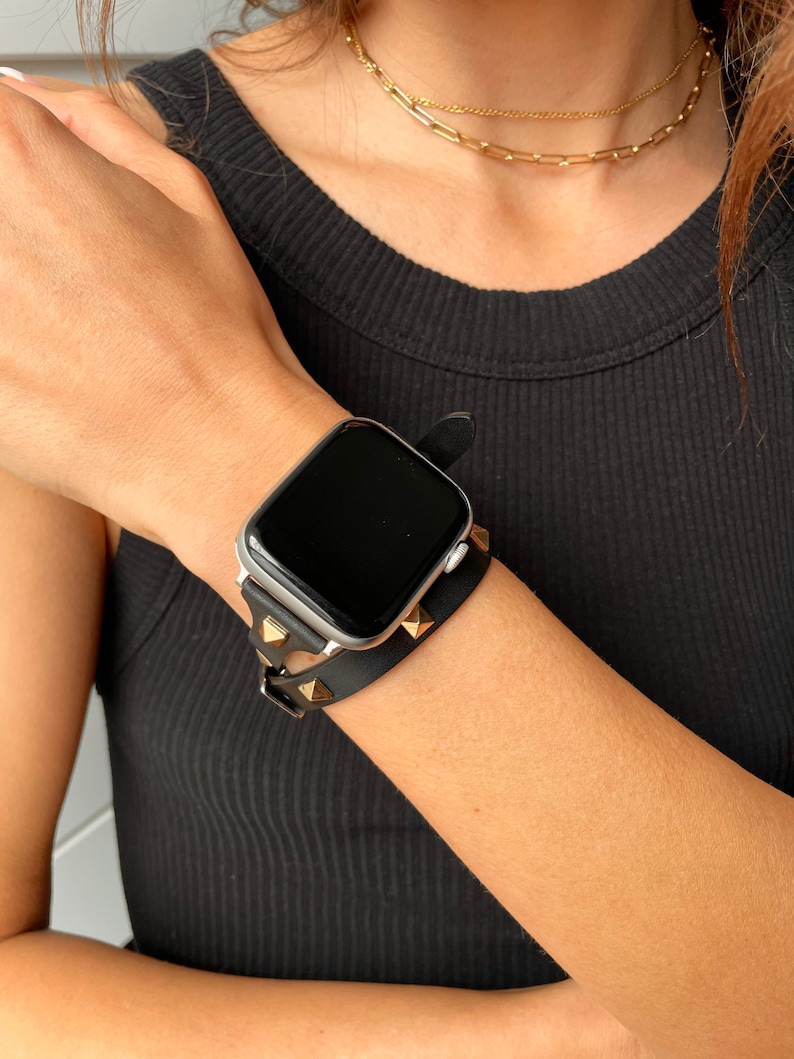 May include: A black leather Apple Watch band with gold studs. The band is double-layered and has a minimalist design.