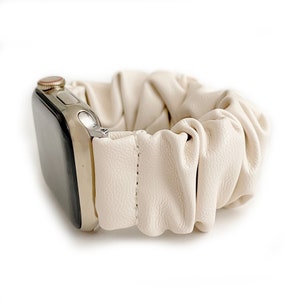 Op de afbeelding: Cr&egrave;me-kleurige leren scrunchie horlogeband. De band is bevestigd aan een zilveren Apple Watch. Het scrunchie-ontwerp is geplooid en getextureerd, met een gestikt naaddetail. De wijzerplaat van het horloge is zwart.