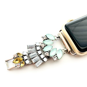Puede incluir: Una pulsera de reloj plateada con una esfera de reloj dorada y un dije decorativo con cristales azul claro, blanco y amarillo.