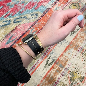 May include: A black leather watch band with gold accents and a gold bangle bracelet with a row of diamonds. The watch band has a studded design. The bracelets are worn on a person's wrist.