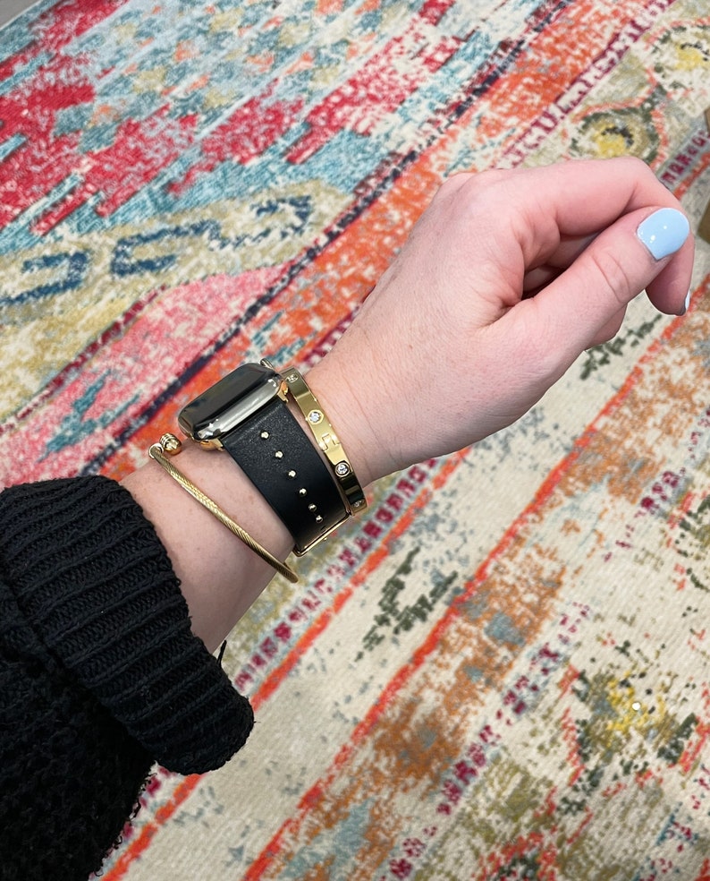 May include: A black leather watch band with gold accents and a gold bangle bracelet with a row of diamonds. The watch band has a studded design. The bracelets are worn on a person's wrist.