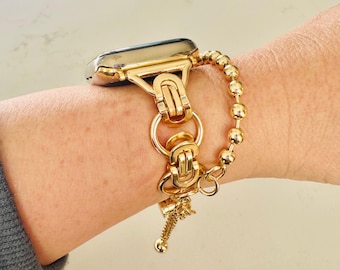 May include: A gold-toned smartwatch band featuring a complex interlocking link design connected to a polished gold-toned watch case, accompanied by a delicate gold-toned beaded chain bracelet that drapes alongside the main watch strap.