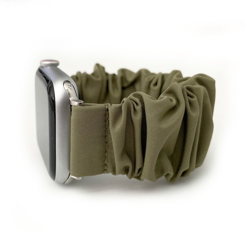 Puede incluir: Una correa de reloj scrunchie verde oliva sujeta a un Apple Watch plateado. La correa est&aacute; hecha de tela fruncida, creando una apariencia texturizada y con volantes. La esfera del reloj es visible. La correa est&aacute; dise&ntilde;ada para un reloj inteligente.