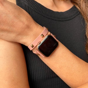 May include: A pink leather Apple Watch band with gold studs. The band is double-wrapped around the wrist.