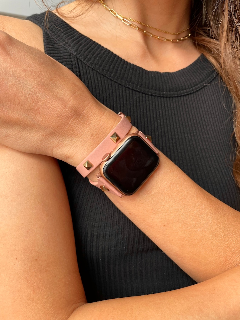 May include: A pink leather Apple Watch band with gold studs. The band is double-wrapped around the wrist.