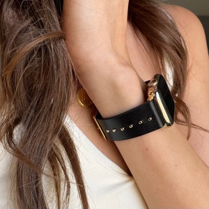 May include: A black leather watch band with gold accents and small gold studs. The band is attached to a gold Apple Watch.