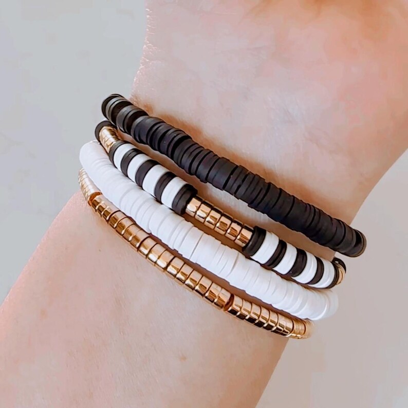 May include: A set of four bracelets featuring black, white, and gold beads. The bracelets are stacked on a person's wrist.
