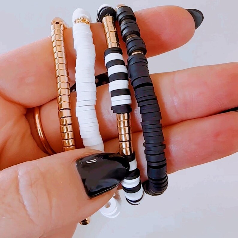 May include: Four bracelets stacked on a hand. The bracelets are made of white and black beads, and one has a gold chain.
