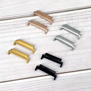 Puede incluir: Ocho cierres de metal para correas de reloj en oro, plata, bronce y negro. Los cierres rectangulares tienen un diseño curvo y están dispuestos sobre una superficie de madera clara. Acabados metálicos.