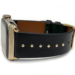 May include: A black leather Apple Watch band with gold accents and studs. The band is shown on a silver Apple Watch.