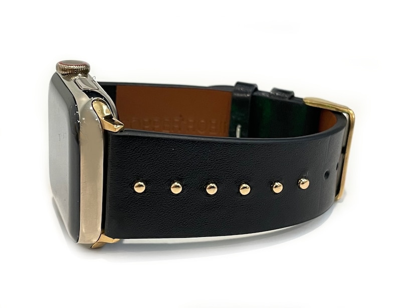 May include: A black leather Apple Watch band with gold accents and studs. The band is shown on a silver Apple Watch.