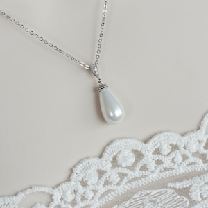 May include: A silver necklace with a pear-shaped, white pendant. The pendant is suspended from a delicate chain and is accented with small, clear stones. The necklace is displayed on a neutral background with white lace trim.