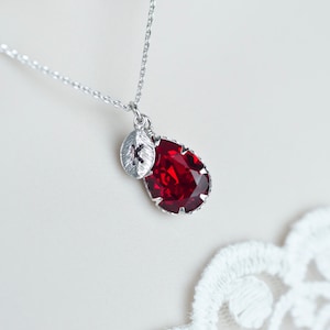 Può includere: Una collana d'argento con un ciondolo di cristallo rosso a forma di lacrima e un piccolo ciondolo d'argento con la lettera "K" su di esso.