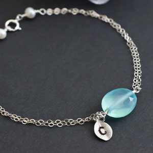 May include: A delicate silver chain bracelet with a light blue gemstone and a silver charm with the letter "C" hanging from it. The bracelet also features two small white pearls.