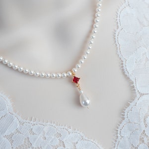 Può includere: Una collana di perle bianche con un ciondolo di pietra rossa e una singola perla a goccia.