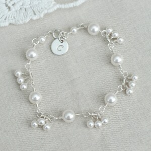 Puede incluir: Un delicado brazalete de plata con un dije con la letra 'C' y un racimo de perlas blancas.