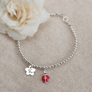 Könnte beinhalten: Ein silbernes Perlenarmband mit einem Glücksbringer in Form eines Marienkäfers und einem silbernen Anhänger mit dem Buchstaben K.