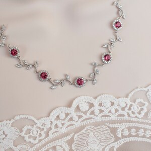Può includere: Una collana d'argento con un delicato design a forma di vite e pietre preziose rosa. La collana è su uno sfondo di pizzo bianco.