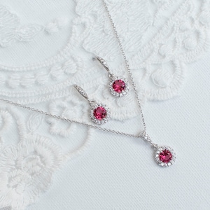 Puede incluir: Un collar y pendientes de plata con una piedra preciosa roja rodeada de piedras preciosas transparentes más pequeñas.