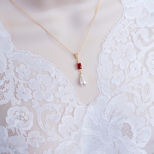 May include: A delicate gold necklace with a red gemstone pendant and a single pearl drop. The necklace is on a white lace background.