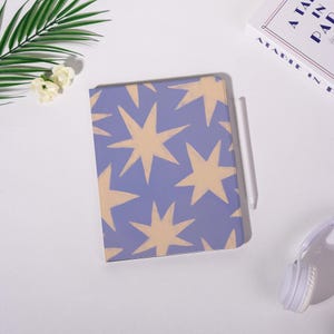 K&ouml;nnte beinhalten: Ein Notizbuch mit blauem Einband und einem Muster aus beigefarbenen Sternen. Ein wei&szlig;er Stift liegt neben dem Notizbuch. Die Szene spielt sich auf einer wei&szlig;en Oberfl&auml;che ab, mit einem Buch und Kopfh&ouml;rern im Hintergrund.
