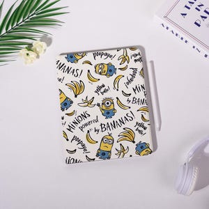 May include: A white tablet case with a repeating pattern of yellow bananas and Minion characters. The case has text that reads "MINIONS powered by BANANAS!" and "yellow bello!" A white stylus rests on the case.