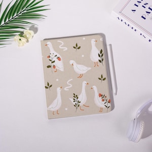 May include: A beige notebook featuring a pattern of white geese with orange beaks and feet, interspersed with green leaves and red berries. A white stylus rests beside the notebook. The notebook is on a white surface.