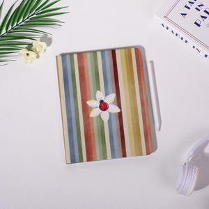 May include: A notebook with a colorful striped pattern in shades of red, yellow, blue, and green. A white flower with a red ladybug in the center is on the cover. A white stylus and headphones are also visible.