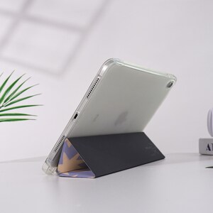 K&ouml;nnte beinhalten: Eine transparente iPad-H&uuml;lle mit einem schwarzen und gemusterten St&auml;nder. Der St&auml;nder ist zu einer dreieckigen Form gefaltet, mit einem Design aus hellvioletten und goldenen Sternformen. Das iPad ist abgewinkelt und zeigt den Bildschirm.