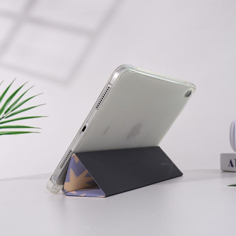 K&ouml;nnte beinhalten: Eine transparente iPad-H&uuml;lle mit einem schwarzen und gemusterten St&auml;nder. Der St&auml;nder ist zu einer dreieckigen Form gefaltet, mit einem Design aus hellvioletten und goldenen Sternformen. Das iPad ist abgewinkelt und zeigt den Bildschirm.