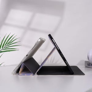 K&ouml;nnte beinhalten: Eine schwarze und graue Tablet-H&uuml;lle mit einem transparenten Tablet im Inneren, das in einem Winkel aufgestellt ist. Die H&uuml;lle ist gefaltet, um einen St&auml;nder zu bilden. Der Hintergrund ist wei&szlig; mit einem Hauch von gr&uuml;ner Pflanze.