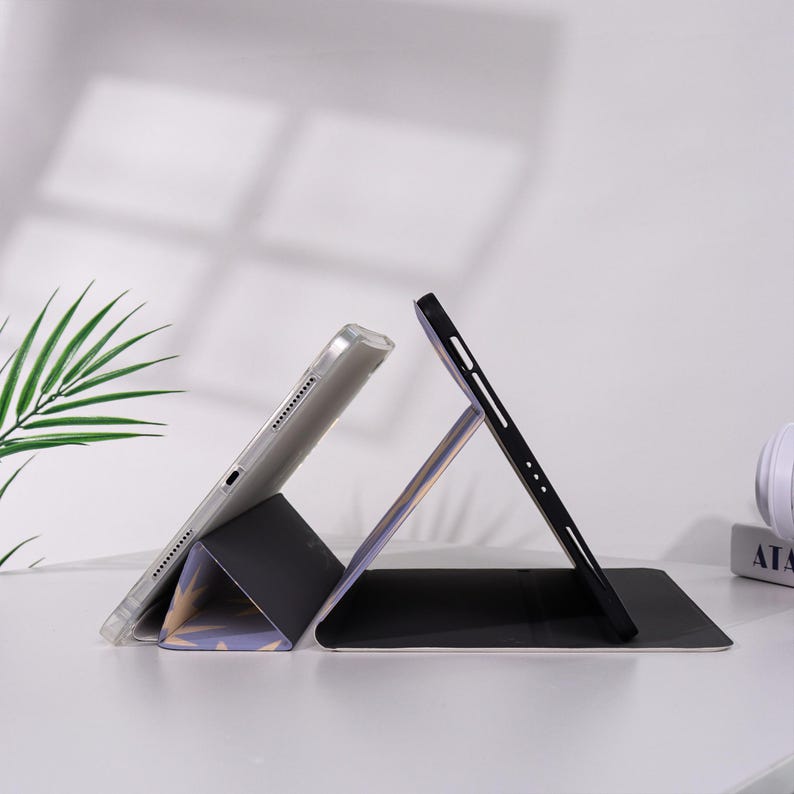 K&ouml;nnte beinhalten: Eine schwarze und graue Tablet-H&uuml;lle mit einem transparenten Tablet im Inneren, das in einem Winkel aufgestellt ist. Die H&uuml;lle ist gefaltet, um einen St&auml;nder zu bilden. Der Hintergrund ist wei&szlig; mit einem Hauch von gr&uuml;ner Pflanze.