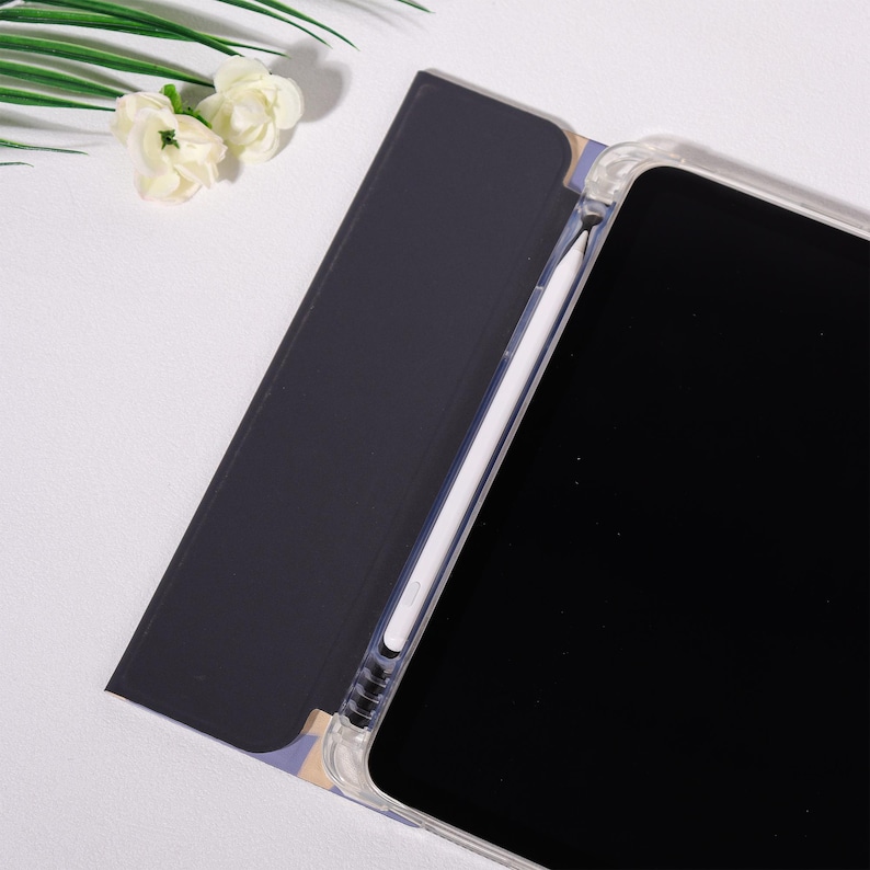 K&ouml;nnte beinhalten: Eine transparente Tableth&uuml;lle mit schwarzem Bildschirm und wei&szlig;em Stift. Die H&uuml;lle ist ge&ouml;ffnet und zeigt eine dunkelblaue Abdeckung mit floralem Muster. Wei&szlig;e Blumen und gr&uuml;ne Bl&auml;tter befinden sich in der oberen linken Ecke.