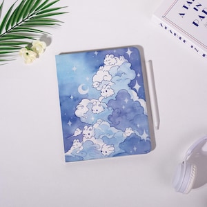 May include: A notebook with a whimsical design featuring fluffy white bunnies in a cloudscape with stars and a crescent moon. The cover is a watercolor wash of blues and purples. A white stylus rests beside the notebook.