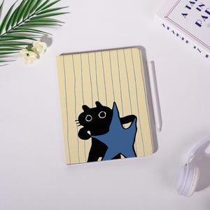 Könnte beinhalten: Ein Notizbuch mit gelb gestreiftem Einband, das eine Illustration einer schwarzen Katze mit einem blauen Stern zeigt. Ein weißer Stift liegt neben dem Notizbuch. Im Hintergrund ein Buch und weiße Kopfhörer.