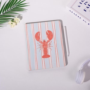 May include: A notebook with a light blue and red striped pattern and a red lobster illustration. A white stylus and headphones are also visible. The notebook is on a white surface.