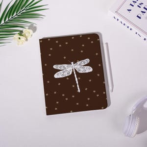 May include: A brown notebook with a white dragonfly design and small white stars. A white stylus rests beside the notebook. The notebook is on a white surface with a book and headphones in the background.