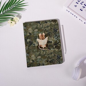 May include: A green and brown patterned notebook featuring a cartoon mouse reading a newspaper. A white stylus and a book titled "A Tale in Paris" are also visible. The notebook has a floral design.