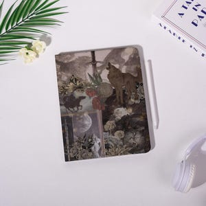 May include: A notebook with a collage design featuring a wolf howling at the moon, a knight on horseback, and floral elements. The cover has a sepia tone with a vintage aesthetic. A white stylus rests on the notebook.