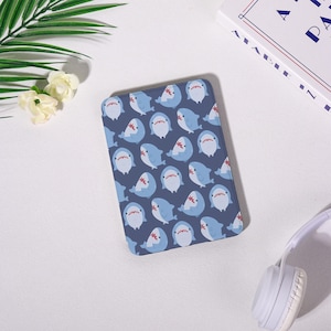 May include: A rectangular, navy blue phone case featuring a repeating pattern of cartoon sharks in light blue and white. The sharks have red mouths and are set against a dark blue background. The case is on a white surface.