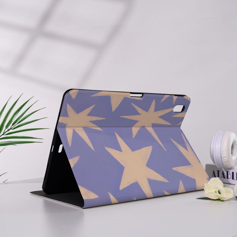 K&ouml;nnte beinhalten: Eine Tablet-H&uuml;lle mit lavendelfarbenem Hintergrund und einem Muster aus gro&szlig;en, tanfarbenen Sternen. Die H&uuml;lle ist ge&ouml;ffnet und zeigt ein schwarzes Innenleben. Das Design ist modern und auff&auml;llig, geeignet zum Schutz eines Tablets.