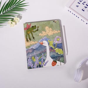 May include: A whimsical notebook featuring a colorful illustration of a goose, a frog fairy, and a mountain landscape. The cover art includes purple flowers, a strawberry, and a stylized sky. A white stylus and a book are also visible.