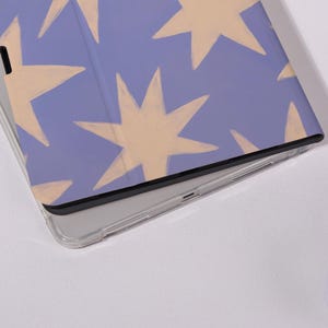 K&ouml;nnte beinhalten: Eine Tablet-H&uuml;lle mit hellviolettem Hintergrund und einem Muster aus gro&szlig;en, cremefarbenen Sternen. Die H&uuml;lle hat eine transparente Schutzh&uuml;lle und einen schwarzen Rand.