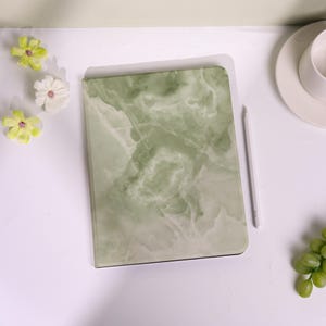 May include: A rectangular tablet case featuring a sage green and white marble pattern with a smooth finish. The case is positioned vertically next to a slim white stylus, accompanied by three small light green flowers and a white ceramic cup.