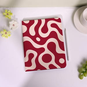 May include: A red notebook featuring a cream-coloured, abstract wavy pattern. A white stylus lies beside the notebook. The notebook is on a white surface, with a cup and saucer in the background.