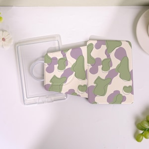 May include: A clear plastic phone case with a matching phone grip and a phone wallet. The phone wallet and grip have a cream-colored background with a green and purple camouflage pattern. The phone case is open.