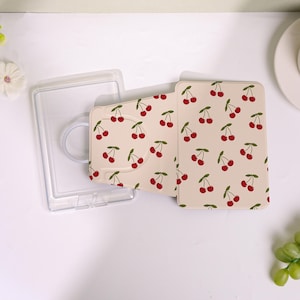 May include: A clear plastic phone case and a beige cardholder with a cherry pattern. The cardholder is open, revealing the interior. The cherries are red with green stems and leaves, set against a light background.