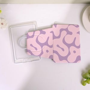 Könnte beinhalten: Eine rosa Handyhülle mit einem abstrakten Lavendelmuster, neben einer transparenten Handyhülle aus Kunststoff. Die Handyhülle ist geöffnet und zeigt die Kamera des Handys. Das Design ist ein sich wiederholendes, wellenförmiges Muster.