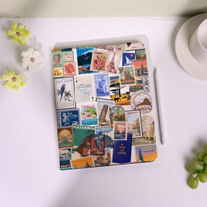 May include: A white tablet case covered in travel-themed postage stamp designs. The stamps feature images of landmarks, landscapes, and text from various locations, including Italy, London, and Los Angeles. A white stylus rests beside the case.