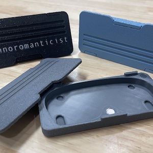 May include: Four 3D-printed rectangular cases in black, gray, and blue. The black case has the word "Technoromanticist" printed on it. The cases have a ridged design and are open or partially open, revealing their interior.